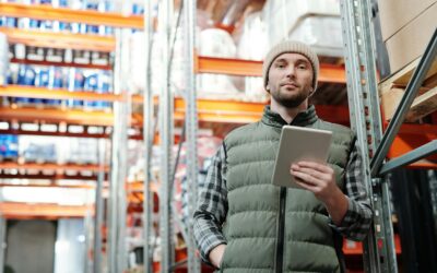 Homem num armazém a segurar um tablet, rodeado por prateleiras com stock, a verificar informação em tempo real.